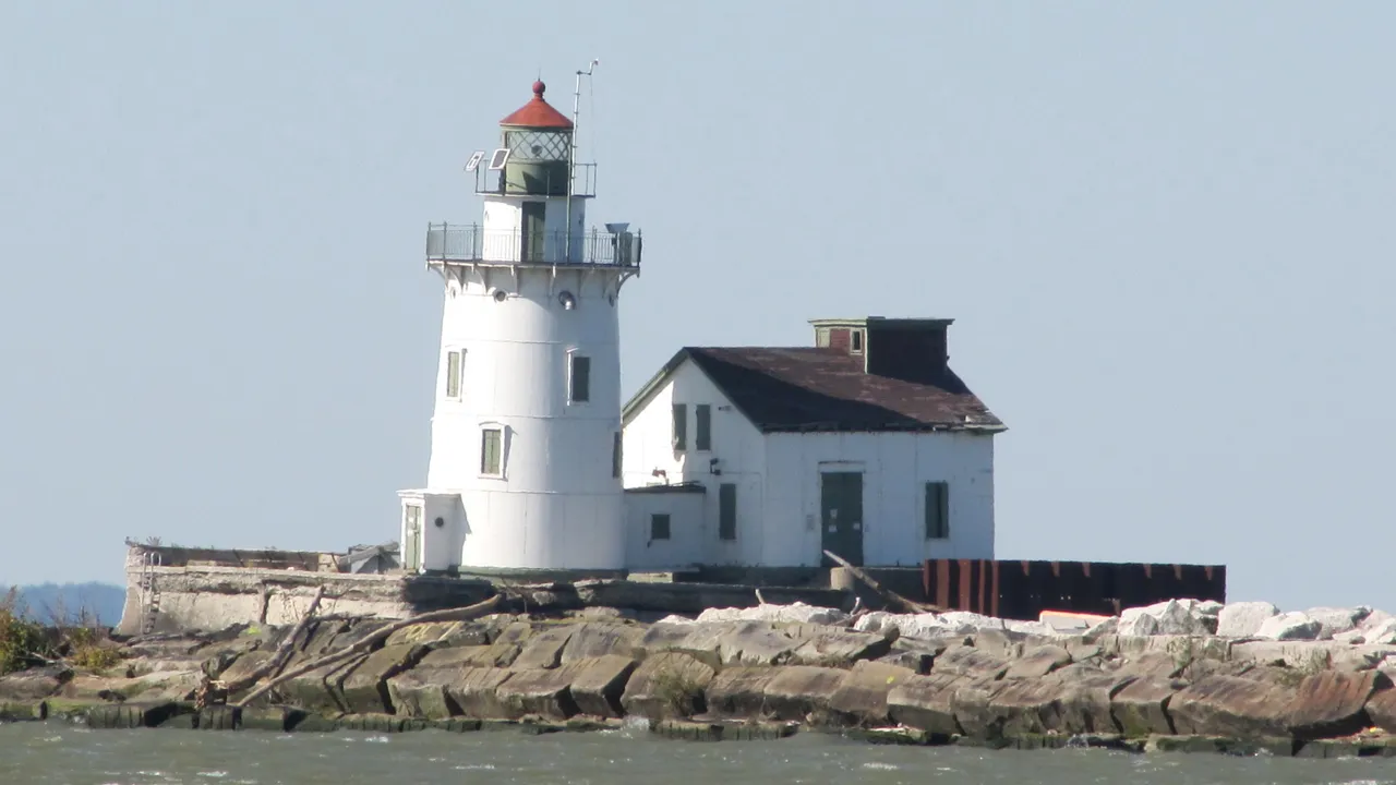 The Cleveland West Pierhead lighthouse, a white tower with a red-roofed lantern beside a white fog house on a stone breakwater under a pale sky.