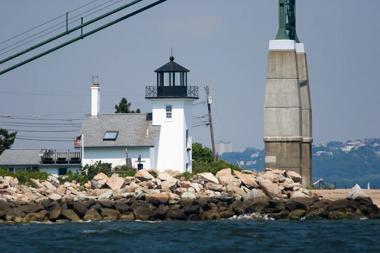 The white Bristol Ferry Lighthouse tower with a black lantern beside its keeper's house, seen from the water over a rock seawall with the Mount Hope Bridge tower beside it.