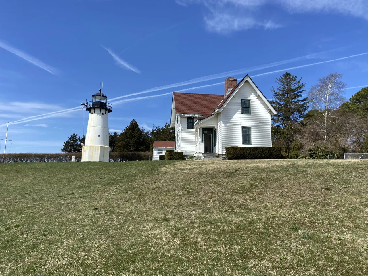 Warwick Neck Lighthouse, a white tower with a black lantern, standing on a grassy rise beside a white two-story keeper's house with a red roof under a blue sky.