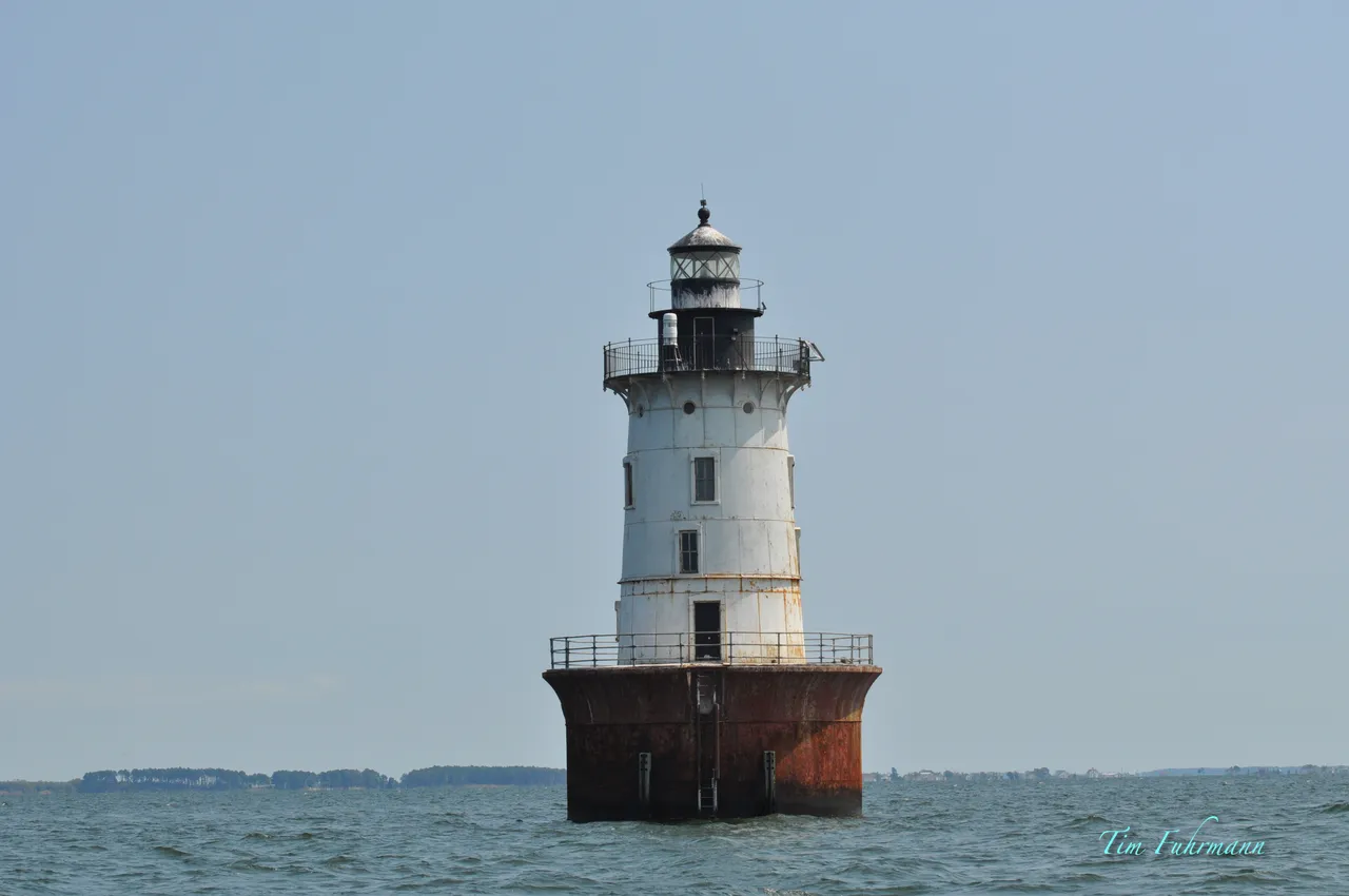 Hooper Island Lighthouse in open water, a white tower with a black lantern on a rust-brown round base under a pale blue sky.