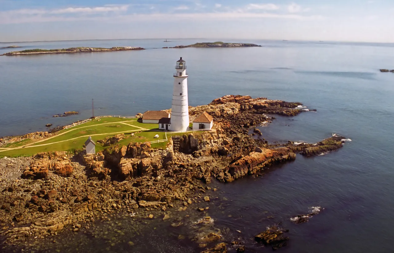 Aerial view of Little Brewster Island with the white Boston Light tower, the keeper's house and outbuildings on green grass above a rocky shoreline.