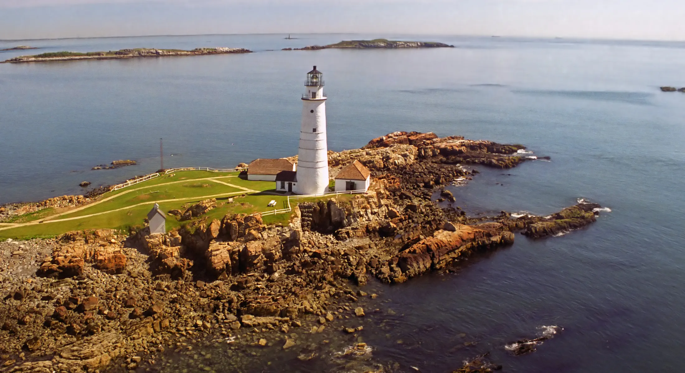 Aerial view of Little Brewster Island in Boston Harbor with the white Boston Light tower, the keeper's house and outbuildings on green grass above a rocky shore.