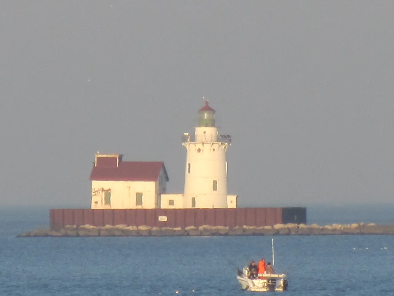The lighthouse and fog house at the end of the breakwater in warm light with a small fishing boat passing in front.