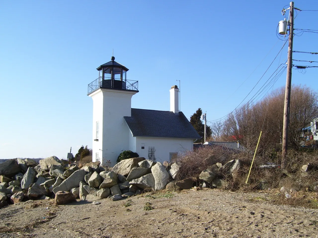 The white lighthouse tower and attached house behind a pile of rocks on a sandy shore under a clear blue sky.