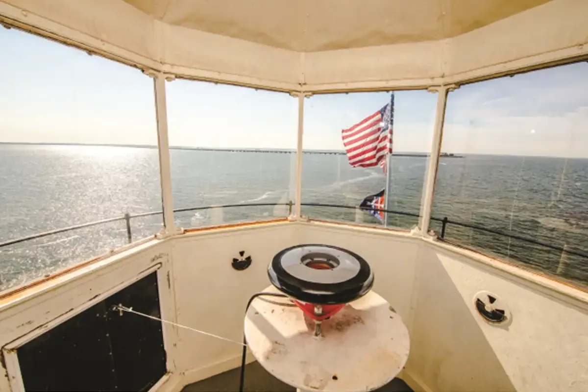 Inside the glassed lantern room looking across the water toward a bridge, with an American flag and the lamp mounted on a white pedestal.