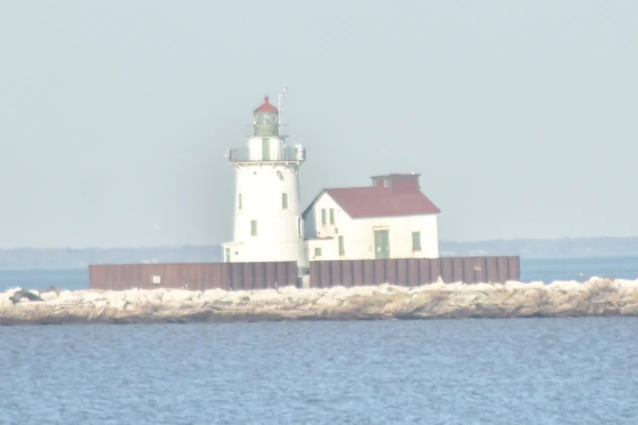 The white tower and red-roofed fog house on a breakwater with ice piled along the stones in the foreground.