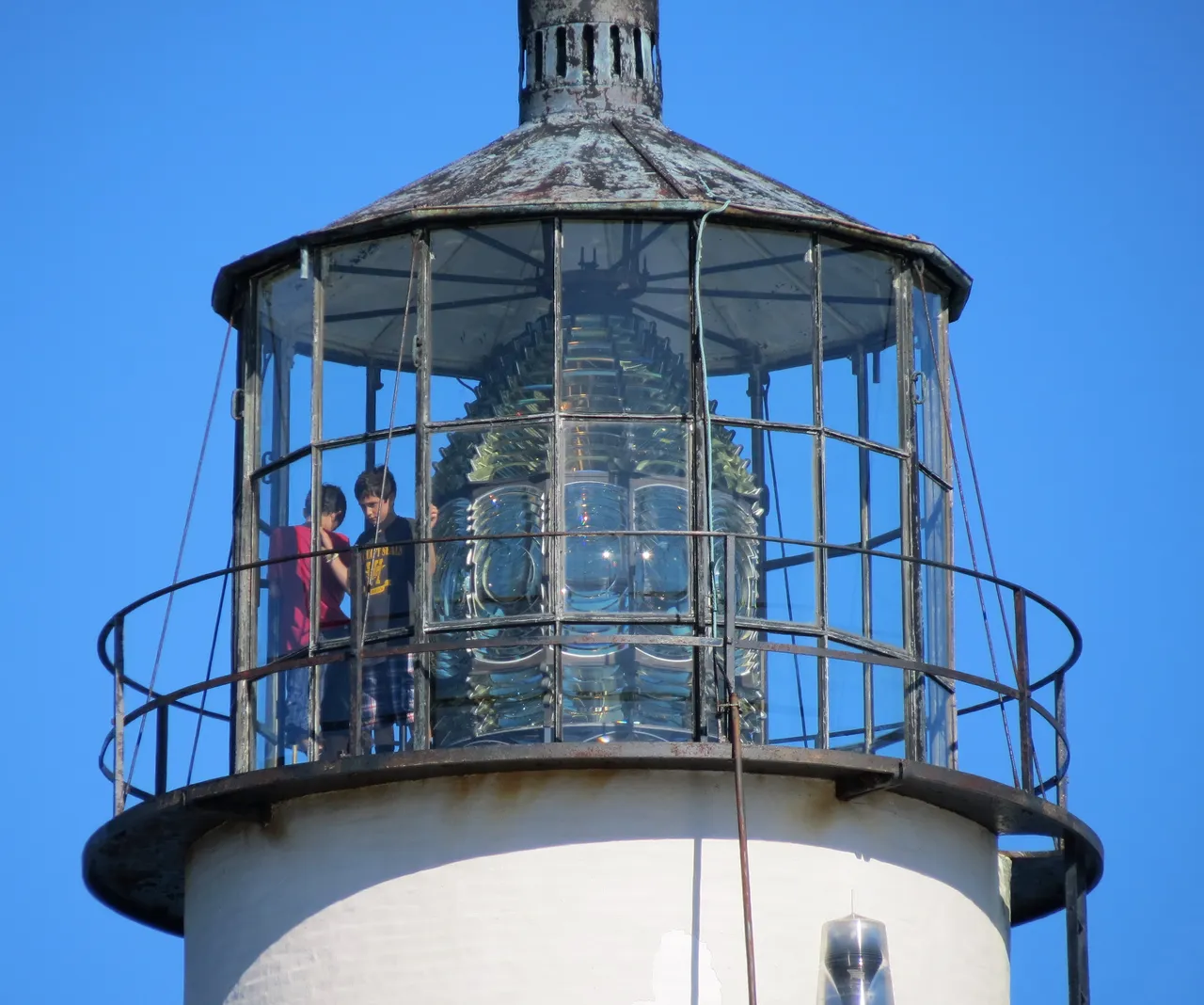 The lantern room and gallery at the top of Boston Light with two people inside beside the large glass lens against a deep blue sky.