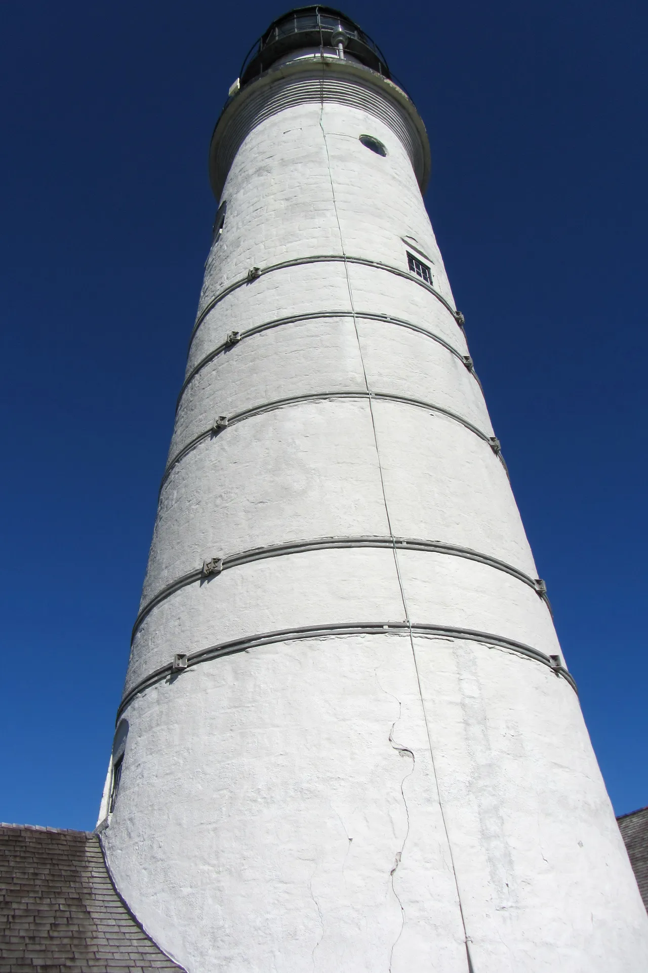 Boston Light's tower seen from below, a tapering white cone with dark iron bands against a blue sky.