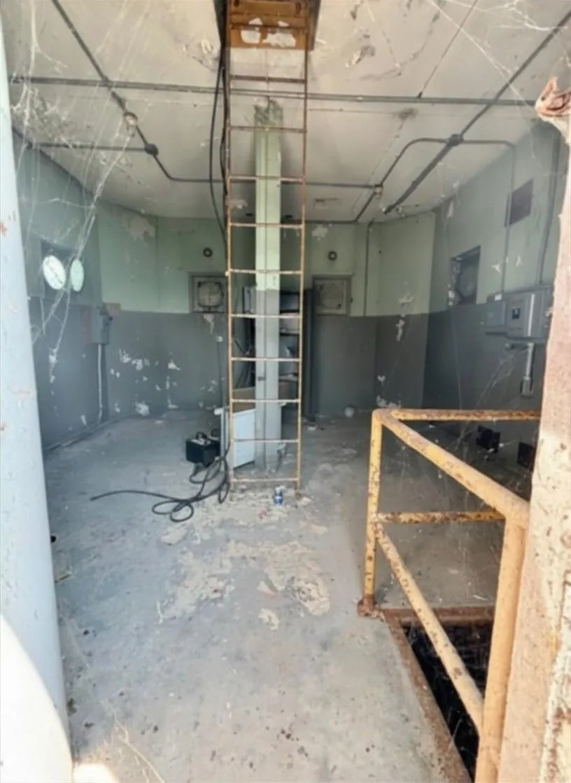 A steel-walled interior room with a ladder and a hatch in the floor inside a lighthouse.
