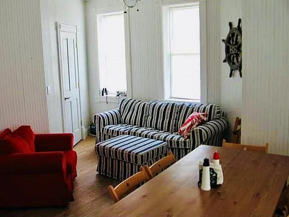 Living area inside the lighthouse with a striped sofa, a red armchair, a wooden table and a ship's wheel on the wall.