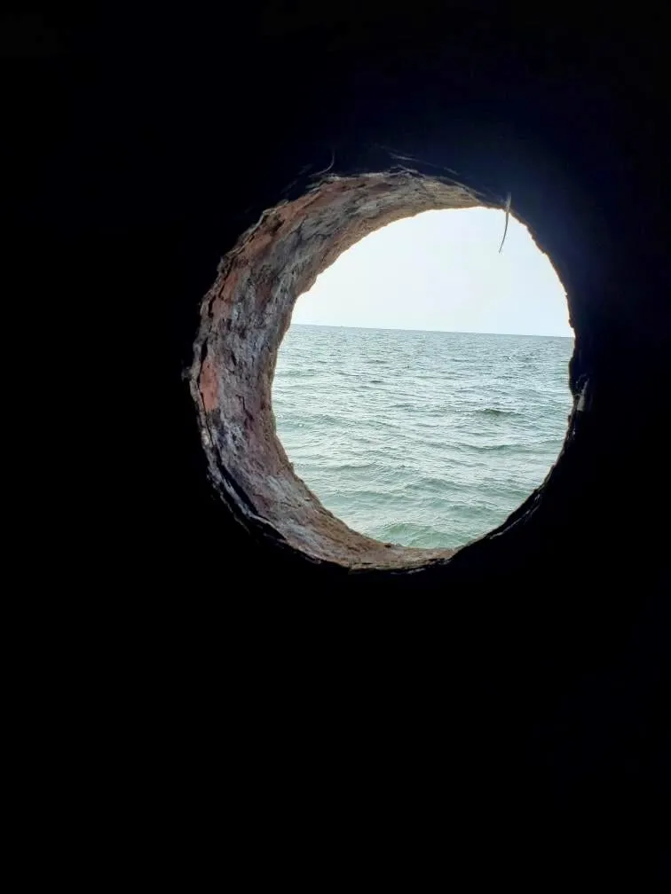 A view of the bay and horizon through a round porthole opening in a dark wall.